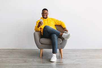 Black Man Using Smartphone Sitting In Armchair Near Gray Wall