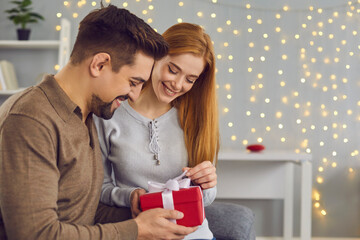 Side view of young married couple unpacking holiday gift together sitting on sofa at home. Man and woman celebrate Christmas or Valentine's Day. People and holidays concept