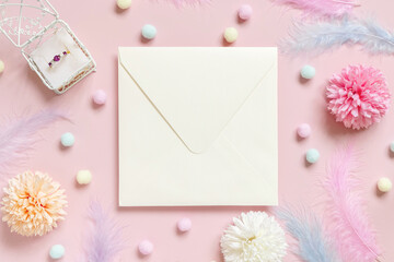Fototapeta na wymiar Square envelope between pastel flowers, pom-poms and feathers near ring in a gift box on pink
