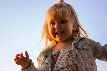 Cute blonde-haired white pre-school girl 2-4 years old in denim jacket waving with hand at camera and smiling. Happy carefree childhood concept. Little child, kid has fun outdoors against blue sky.