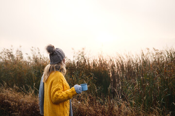 Woman relaxing with coffee next to lakeshore at misty cold morning. Camping and hiking outdoors in nature. Blond hair woman wearing knit hat and sweater