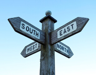 Wooden directional sign post displaying the compass points North, East, South, West. Set against a clear, pale blue sky - Powered by Adobe
