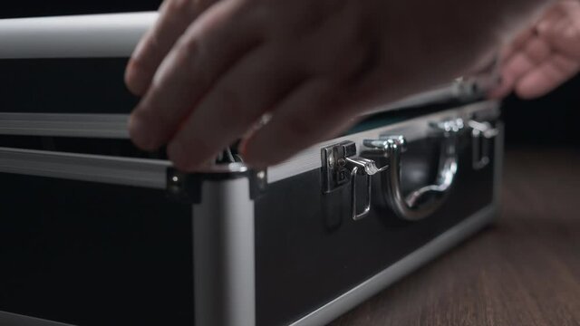 The man carefully opens the case for storing and transporting studio microphones. Aluminum case with locks. Close-up shooting. Black background.