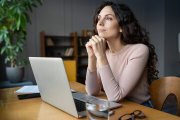 Dreamy businesswoman freelance writer looking in window, distracted from laptop, sitting at work desk in office. Happy young female freelancer thinking of new opportunity, project strategy or vacation