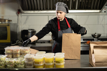 woman wearing an apron and gloves puts disposable plastic food containers into a disposable paper...