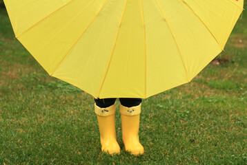 Happy little child, adorable blonde curly toddler male wearing green waterproof coat and yellow boots holding yellow umbrella playing in the garden or park on a sunny rainy warm early autumn day.