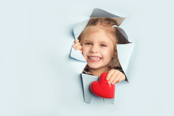 Adorable little girl peeking from ripped paper hole and holding red heart. Valentines, Mother day, birthday or celebration and love concept.
