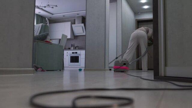 Middle aged woman using vacuum cleaner with long black cord cleaning wooden floor at home