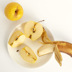 Shriveled yellow apples and overripe banana on a white background. One apple is cut into three pieces. The banana peel is ajar. View from above. Ugly food concept.
