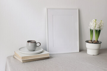 Spring breakfast still life scene. Cup of coffee, books. Empty white picture frame mockup. Beige linen tablecloth. Potted white hyacinth plant. Farmhouse, Scandinavian interior. Easter holiday concept