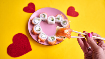 Sushi rolls with salmon in a heart shape and a woman's hand with chopsticks on a yellow background.Creative sushi concept for Valentines day or romantic dinner     