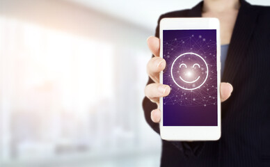 Business satisfaction survey. Hand hold white smartphone with digital hologram Smiley Face sign on light blurred background. Smiley Face Rating for a Satisfaction Survey