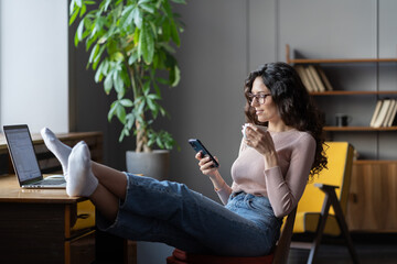 Young calm female entrepreneur relaxing at cozy workplace with legs on table, using mobile phone...