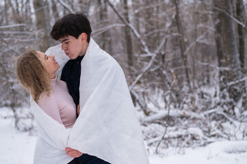 A young couple walks in the park in winter. The guy and the girl are kissing wrapped in a white blanket outdoors.