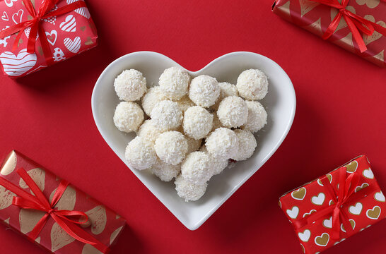 Sweet coconut rafaello candies in a heart-shaped plate and gifts on red background. View from above