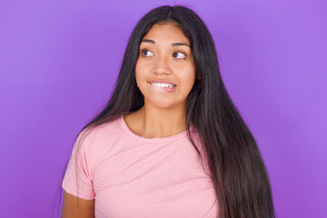 Fototapeta premium Amazed Hispanic brunette girl wearing pink t-shirt over purple background bitting lip and looking tricky to empty space.