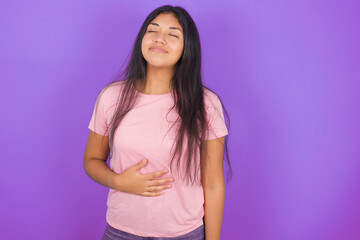 Hispanic brunette girl wearing pink t-shirt over purple background touches tummy, smiles gently, eating and satisfaction concept.