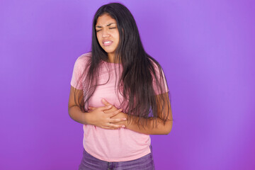 Hispanic brunette girl wearing pink t-shirt over purple background with hand on stomach because nausea, painful disease feeling unwell. Ache concept.
