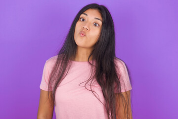 Hispanic brunette girl wearing pink t-shirt over purple background making fish face with lips, crazy and comical gesture. Funny expression.