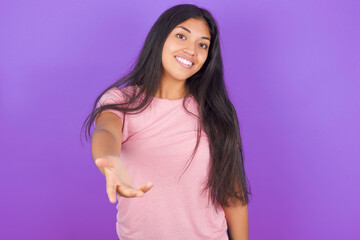 Hispanic brunette girl wearing pink t-shirt over purple background smiling friendly offering handshake as greeting and welcoming. Successful business.