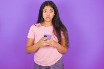Shocked Hispanic brunette girl wearing pink t-shirt over purple background opens mouth hold phone reading advert unbelievable big shopping prices