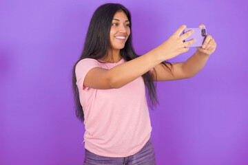 Hispanic brunette girl wearing pink t-shirt over purple background taking a selfie to post it on social media or having a video call with friends.