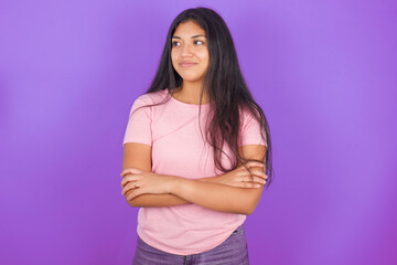 Pleased Hispanic brunette girl wearing pink t-shirt over purple background keeps hands crossed over...