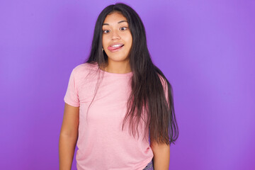 Funny Hispanic brunette girl wearing pink t-shirt over purple background makes grimace and crosses eyes plays fool has fun alone sticks out tongue.