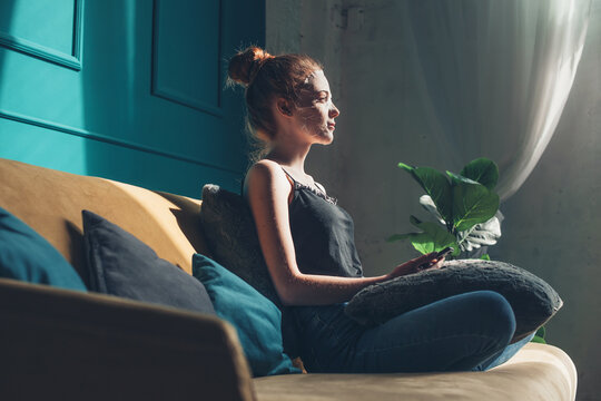 Side view of a woman sitting on sofa in facial mask holding phone. Beauty face. Beauty skin care. Side view.