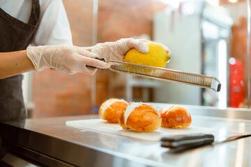 Woman grates fresh lemon zest above buns with cream at metal table