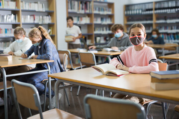 Portrait of preteen girl in protective mask reading books in college library