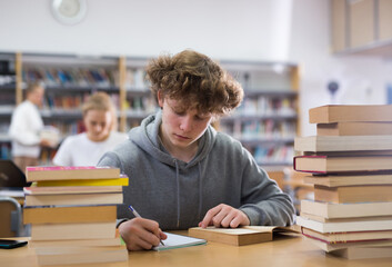 Focused fifteen-year-old schoolboy sitting at a desk in the library is writing an synopsis from a textbook