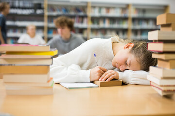 Tired teenager girl sleeping on table among books in library.