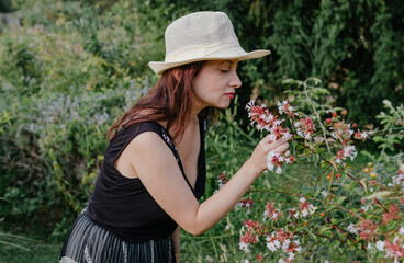 A gorgeous dyed red hair woman with hat smelling flowers in a garden