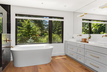 Beautiful bathroom in new luxury home with large soaker bathtub, double vanity with hardwood cabinets and quartz counters. Faucets are mounted to wall. Large window with view of trees.