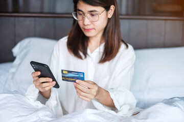 woman using mobile smart phone and credit card for online shopping while making order on bed in morning at home. technology, ecommerce, digital banking online payment and apartment living concept