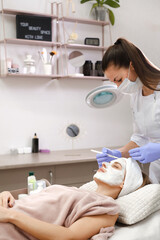 Obraz na płótnie Canvas Cosmetologist apply skin mask with brush, care and treatment procedure in modern aesthetic clinic. Woman treats skin in beauty salon, anti-age non-surgical care for face and natural beauty
