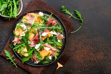 Summer salad with melon, jamon, white cheese and arugula on brown table background, top view, copy space