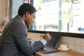 a man working with laptop and writing note,                              african american businessman sitting and focus on his work at coffee bar near glass window indoor cafe