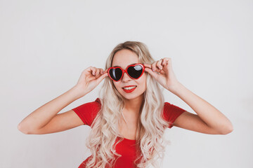 Adorable young blonde girl in red bodysuit and glasses in the shape of hearts posing on white background