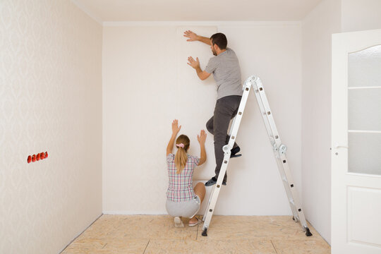 Young adult couple making interior change. Man standing on metal ladder and woman help applying new wallpaper on white wall in room. Working together. Repair work of home. Back view.