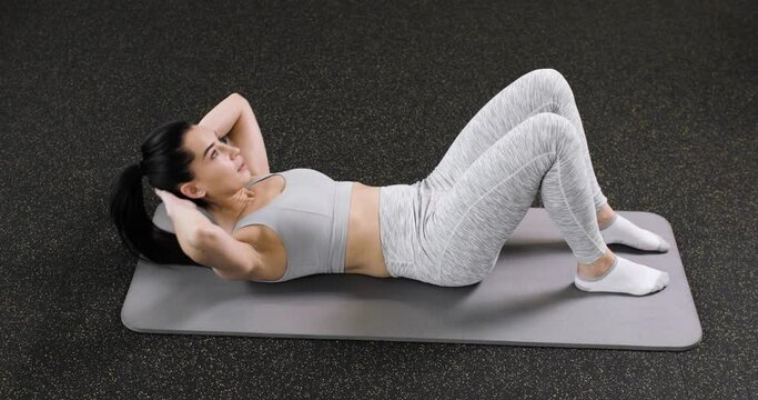 A young caucasian athletic woman does an exercise on the abdominal press, lies on the floor in the gym, dressed in gray sportswear. Side view. Active lifestyle concept.