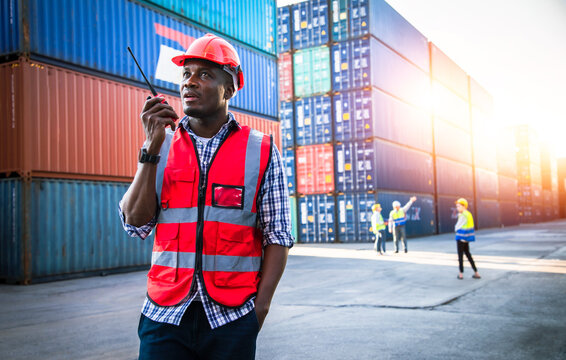Portrait of handsome Africa American engineering using walkie talkie for control labor in front of container store. Back view of assistant on background of cargo store. Export and transportation