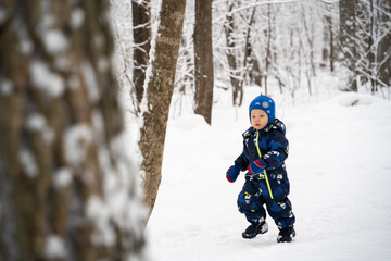 Fototapeta na wymiar Joyful curious and independent toddler kid walks along the path in the winter forest. A little boy in warm overalls and a hat enjoys a walk in the winter snowy forest during winter holidays.
