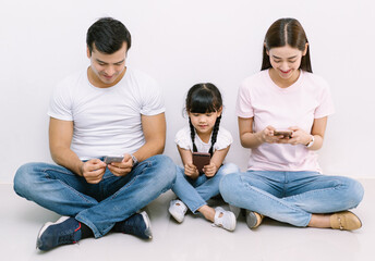 Happy technology addicted Asian family parents and kid holding smart phones having electronic devices sit on floor white background. Gadgets dependence overuse, Social media addiction concept