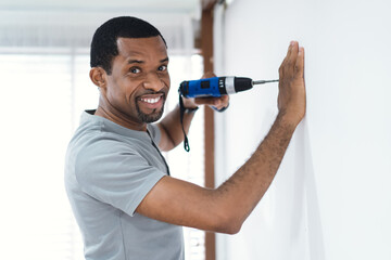 Happy African American man builder drilling a hole in a new white wall in new house with a battery operated hand drill and looking at camera. Adult make DIY workshop. Moving in new house concept - obrazy, fototapety, plakaty