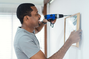 Smiling black man builder drilling a hole in white wall and putting picture frame at home. Home...
