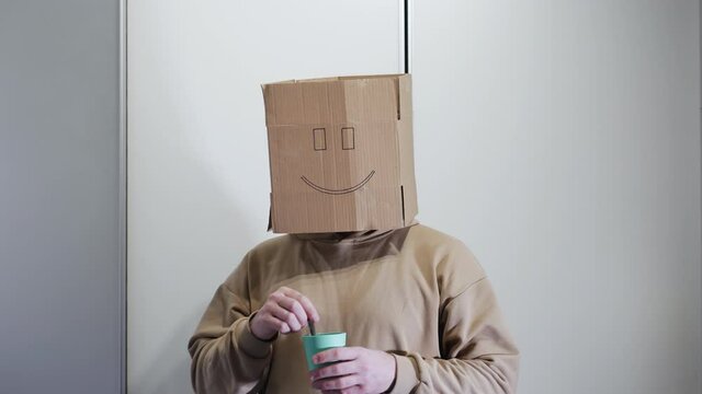 A man with a smile holds a glass with a drink, a funny man stir the drink with a spoon and smiles. A man with a box on his head
