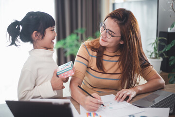 Happy Mother's day. Young woman mom getting congratulations from excited cute little daughter at home, child giving mother gift box while she working on laptop. Family holidays concept