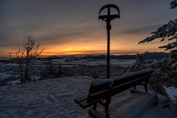 Empty lonely bench and cross with Jesus Christ in cold snowy winter morning in village Martincek, Slovakia